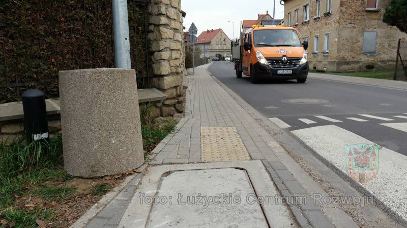 betonowy kosz na śmieci stojący przy chodniku, ulicą jedzie pomarańczowa ciężarówka z logo ZGiUK